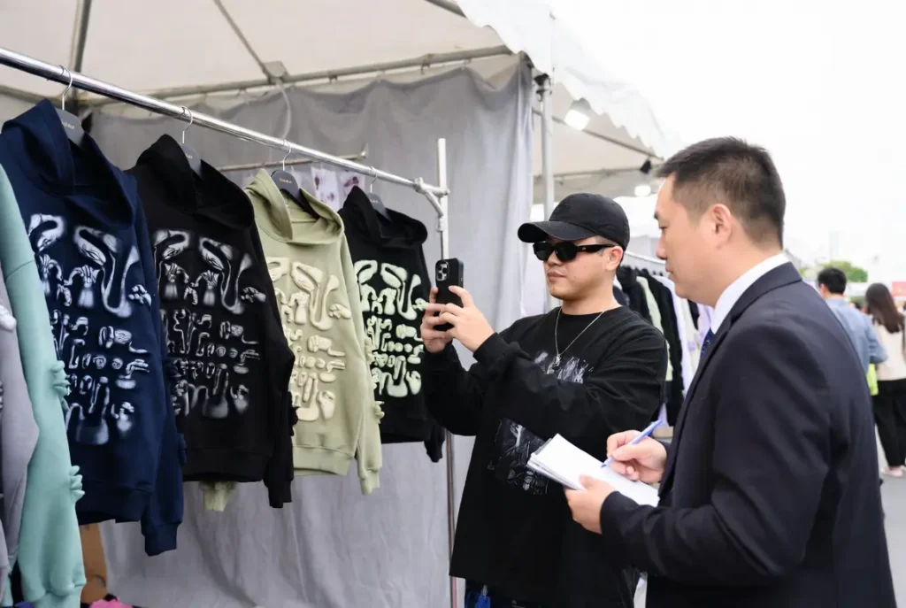 an engaged buyer capturing streetwear samples and notes at a manufacturer booth – exemplifies on-site evaluation and content creation.